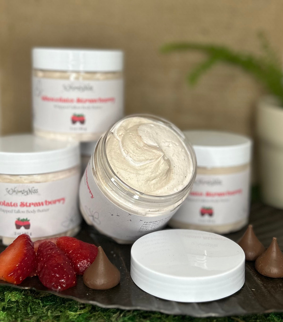 A jar of WhimsyNiss chocolate strawberry scented whipped tallow surrounded by several other jars of the same product. Sitting on a brown shiny try with some fresh strawberries and chocolate kisses.