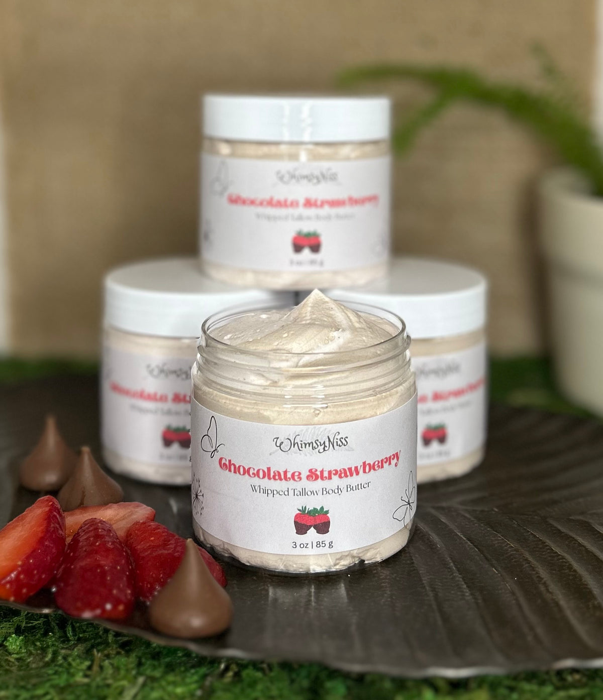 Three jars of WhimsyNiss Chocolate Strawberry Whipped Tallow Body Butter are displayed on a dark textured tray with fresh strawberries, chocolate kisses, and green moss. The front jar is open, showing the whipped body butter, while the other jars are stacked behind it.
