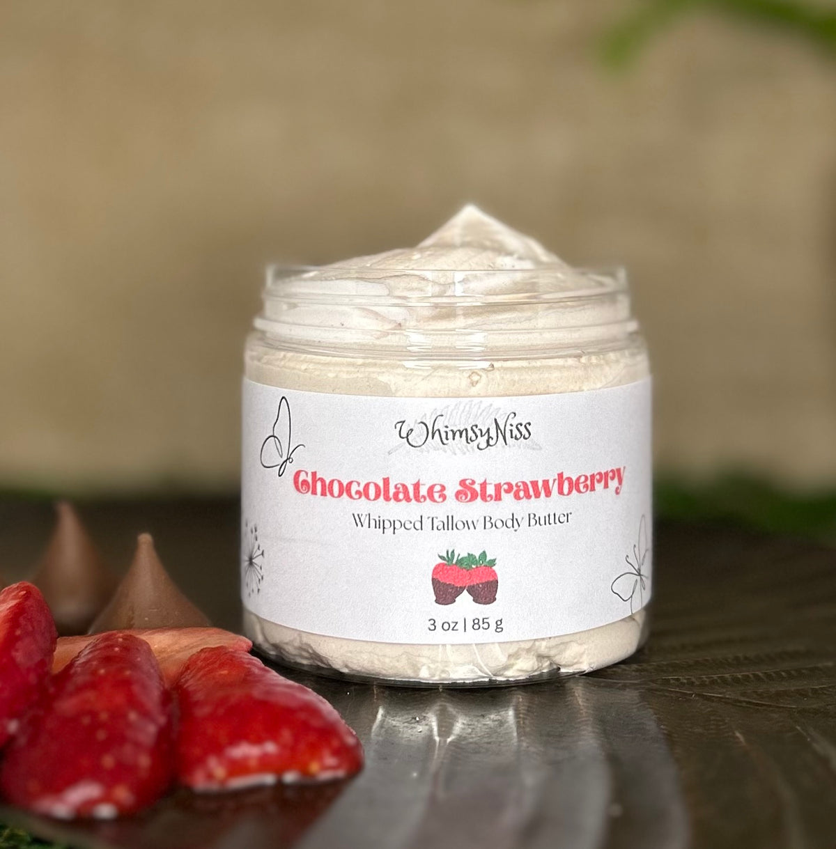 A jar of WhimsyNiss Chocolate Strawberry Whipped Tallow Body Butter sits on a dark reflective surface with fresh strawberries and chocolate kisses in the foreground, against a soft beige background.