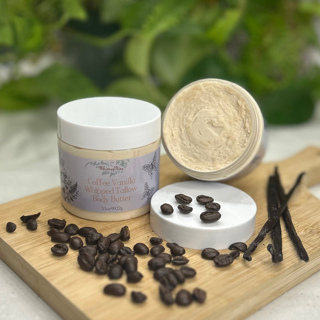 A jar of whipped body butter with its lid off, displayed on a wooden board with coffee beans, vanilla beans, and a blurred green plant background.