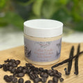 A jar of WhimsyNiss Body Butters Coffee Vanilla Whipped Tallow Body Butter sits on a wooden board surrounded by coffee beans and vanilla beans, with a soft-focus green garden background.