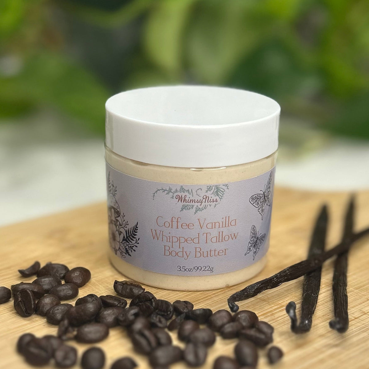 A jar of WhimsyNiss Body Butters Coffee Vanilla Whipped Tallow Body Butter sits on a wooden board surrounded by coffee beans and vanilla beans, with a soft-focus green garden background.