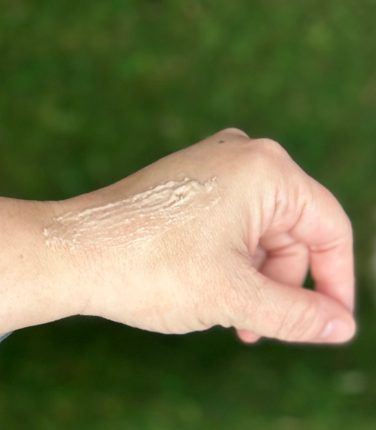 A close-up of a person's hand showing a thick, creamy white lotion or whipped product swatched across the back of the hand, with a soft-focus green outdoor background.