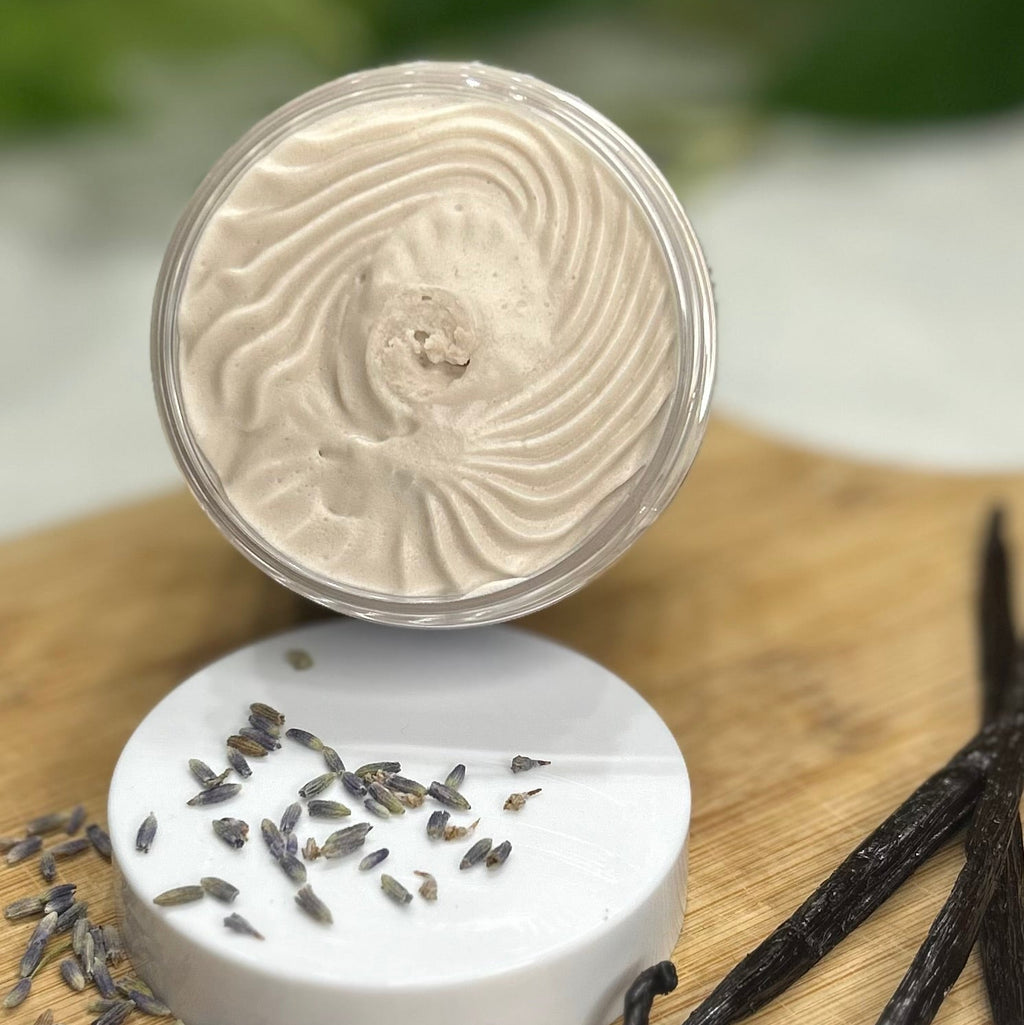 Close-up of an open container of Lavender Vanilla Whipped Body Butter leaning on a lid, surrounded by lavender flowers and vanilla beans on a wooden surface.