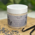 Jar of lavender vanilla whipped tallow body butter with lavender flowers and vanilla beans on a wooden surface, with a blurred background.