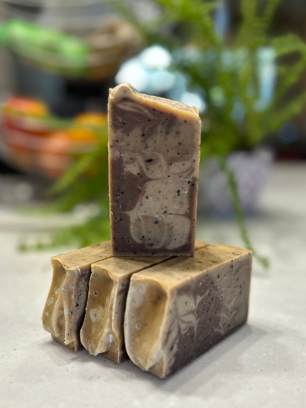 Bar of natural soap on top of 3 lying down soap bars on a white surface with a blurred plant background.